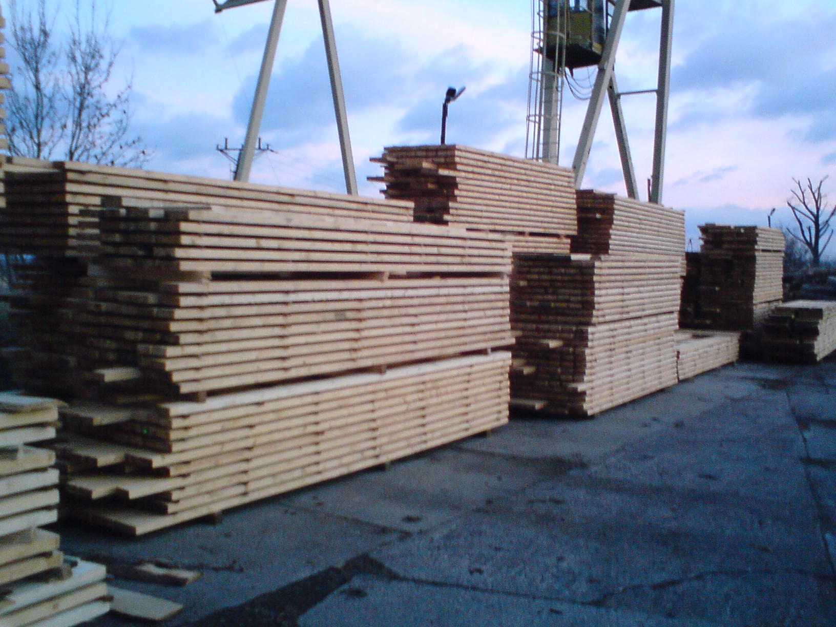 Stosy tarcicy konstrukcyjnej na zewnątrz, widoczne dźwig i pochmurne niebo w tle.